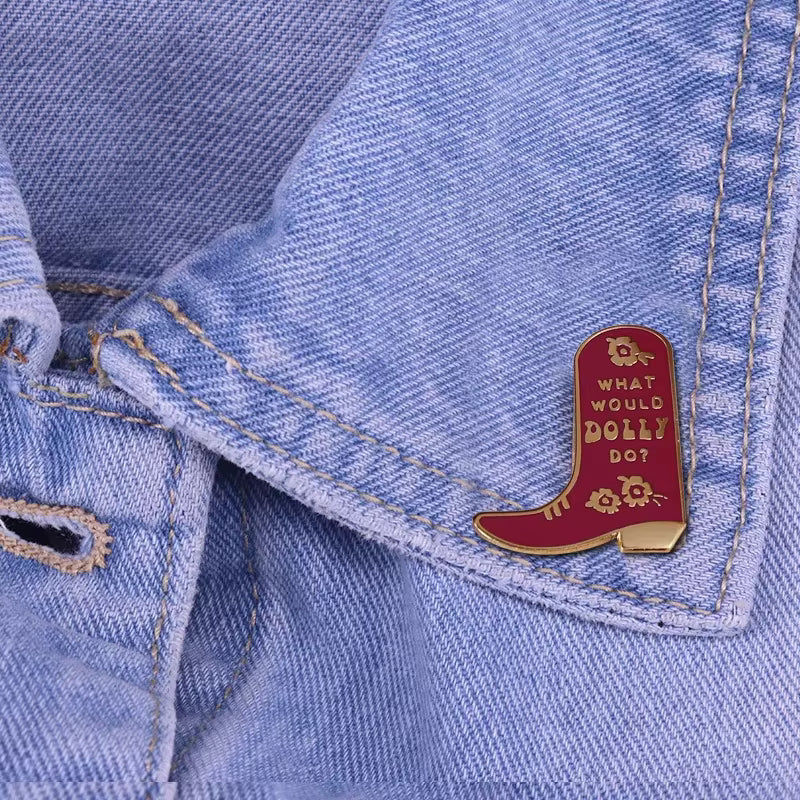 A pink cowboy boot-shaped enamel pin with gold lettering that reads 'WHAT WOULD DOLLY DOT' and small gold floral details, pinned to a light blue denim jacket.