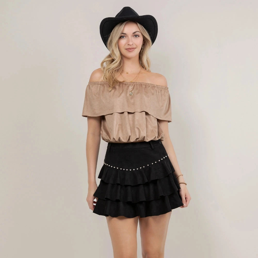 A woman modeling a western-style outfit consisting of a beige off-the-shoulder ruffled top, a black tiered mini skirt with silver studs, a black cowboy hat, and black cowboy boots, against a plain light gray studio background.