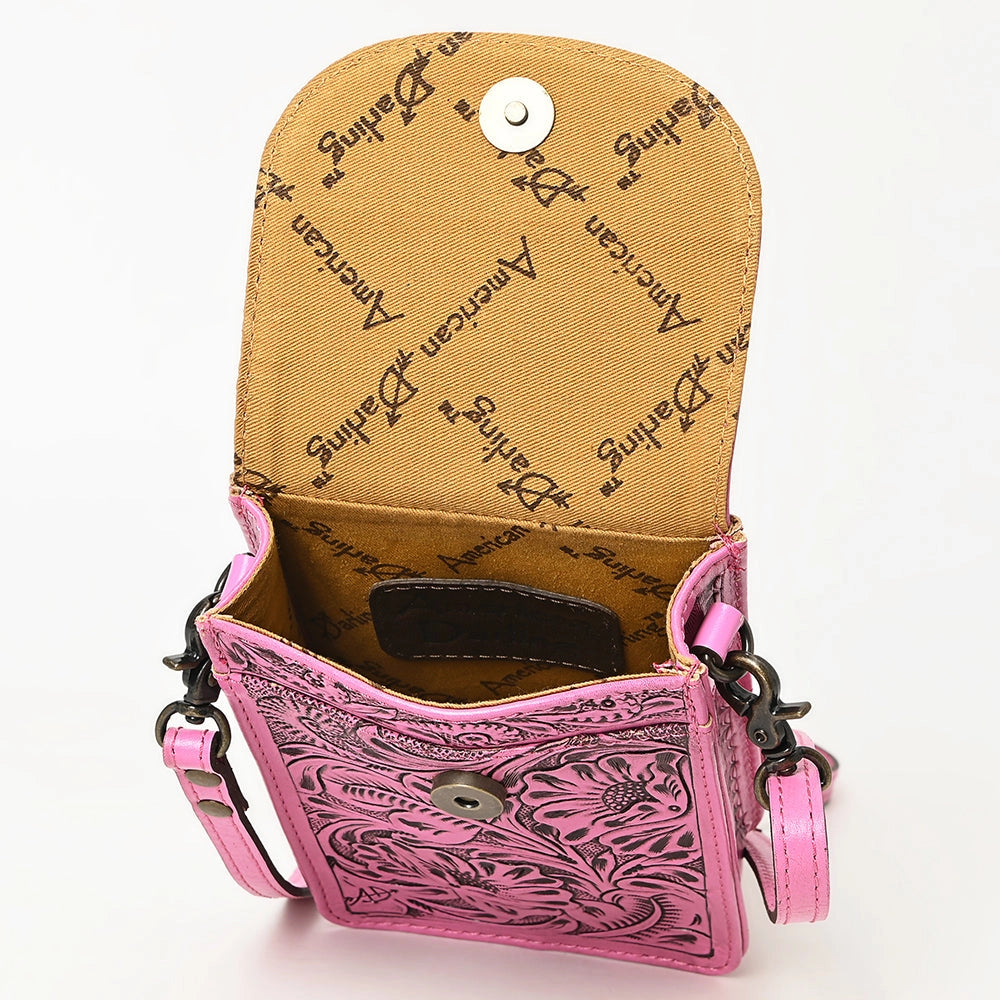 A pink tooled leather crossbody bag with floral embossing, shown open to reveal a tan interior lined with 'American Darling' script. It features a silver snap button closure and a matching pink leather strap.
