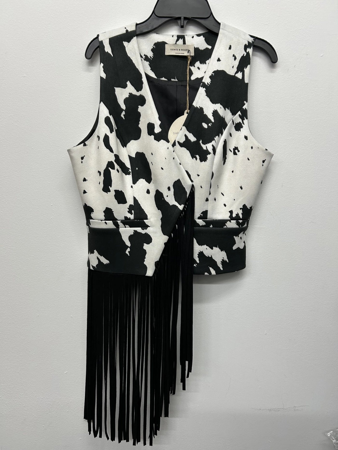 Black and white cow print patterned vest with fringe on a hanger against a light gray background