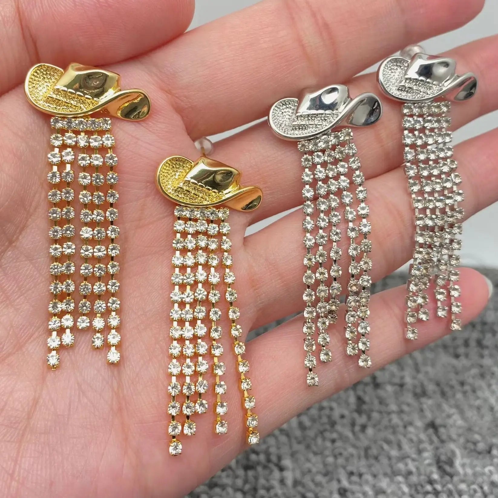 Two pairs of dangling earrings shaped like cowboy hats, displayed on a person's hand. One gold-toned and one is silver-toned, with each hat embellished with clear rhinestones and long cascading rows of rhinestones.