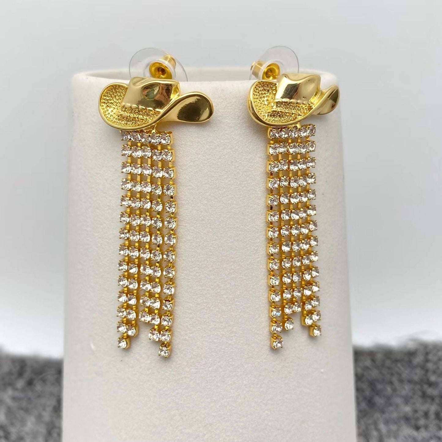 Gold cowboy hat fringe earrings on a white stand with a grey background