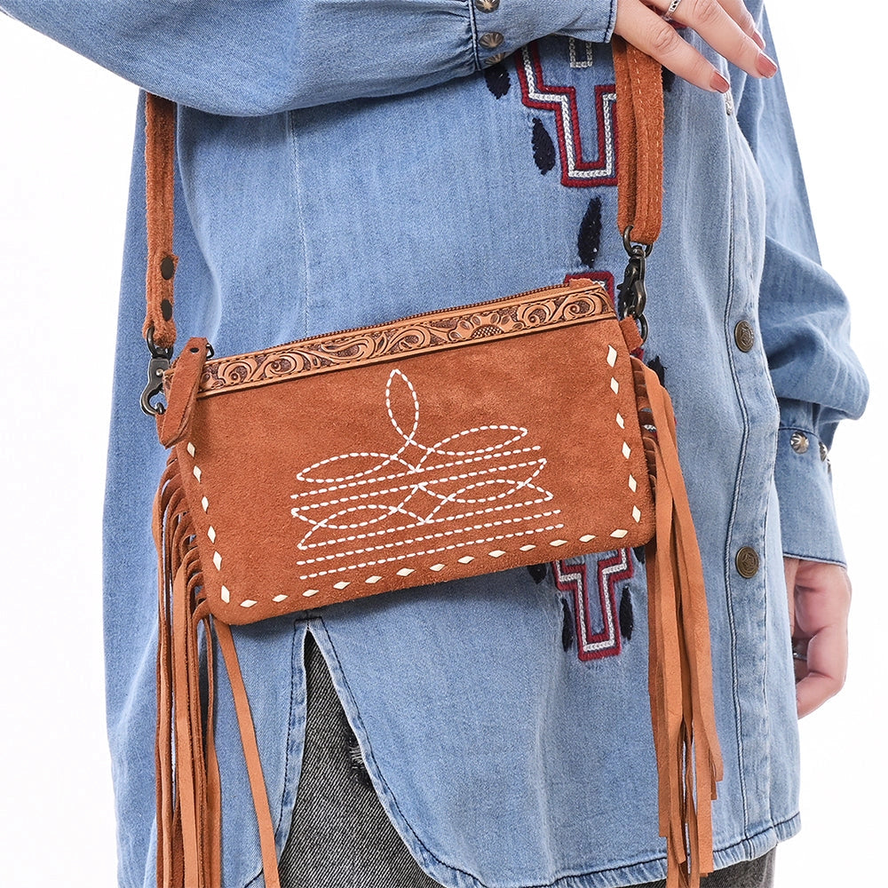 Brown leather crossbody bag with fringe details held by a person wearing a denim shirt.