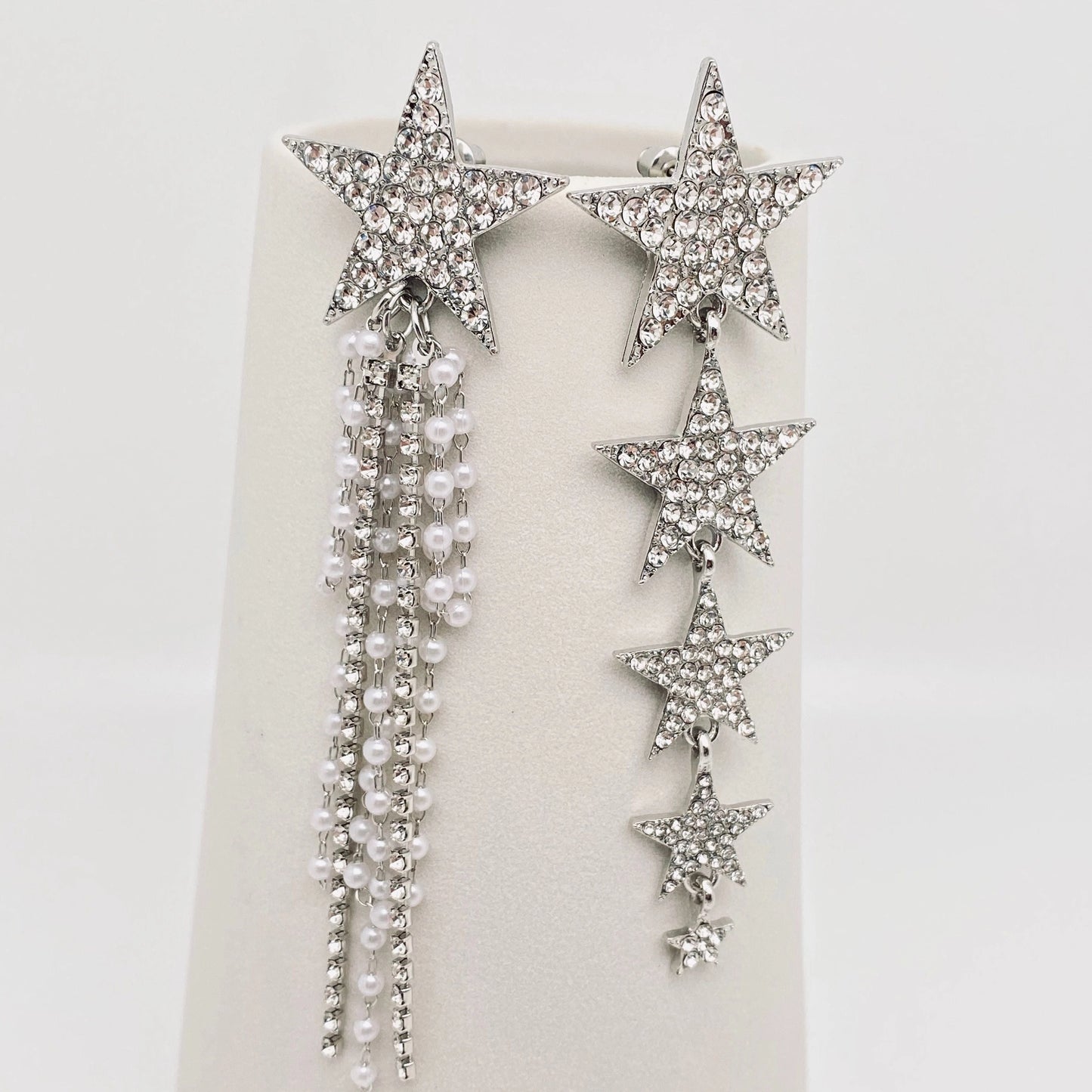 A pair of silver-toned star-shaped drop earrings embellished with clear rhinestones and adorned with dangling strands of white faux pearls, displayed on a cream jewellery stand against a white background.