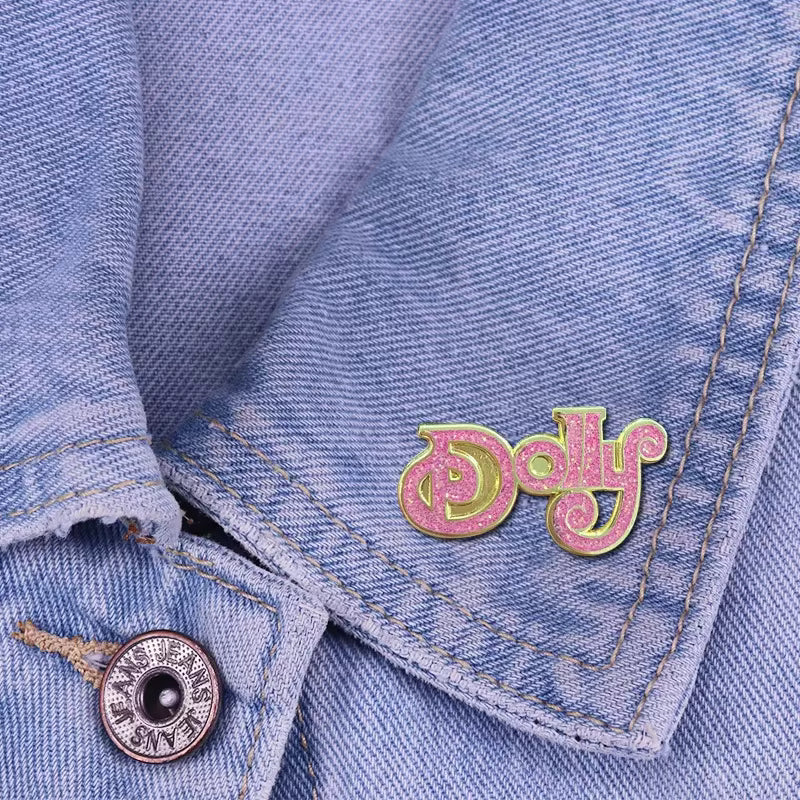 A close-up of a light blue denim jacket showing the collar and button, with a pink glittery enamel lapel pin that reads 'Dolly' in script.