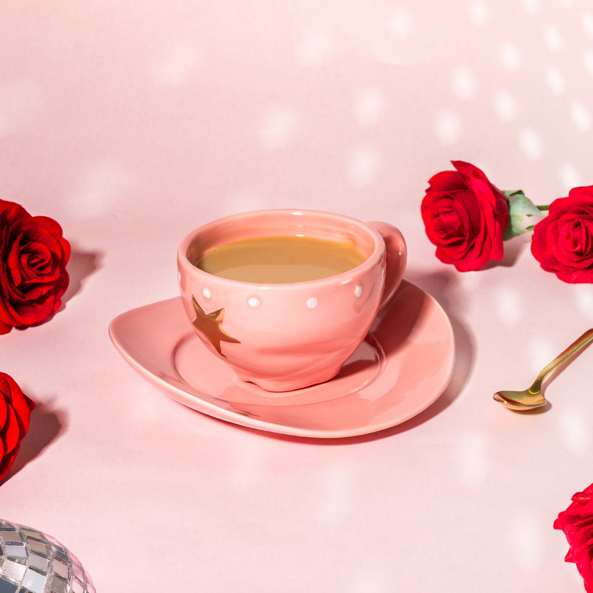 A pink ceramic teacup and saucer set on a pink surface, styled with red roses, a gold spoon, and a disco ball edge. The teacup has white polka dots and a gold star decoration, and contains a light brown beverage.