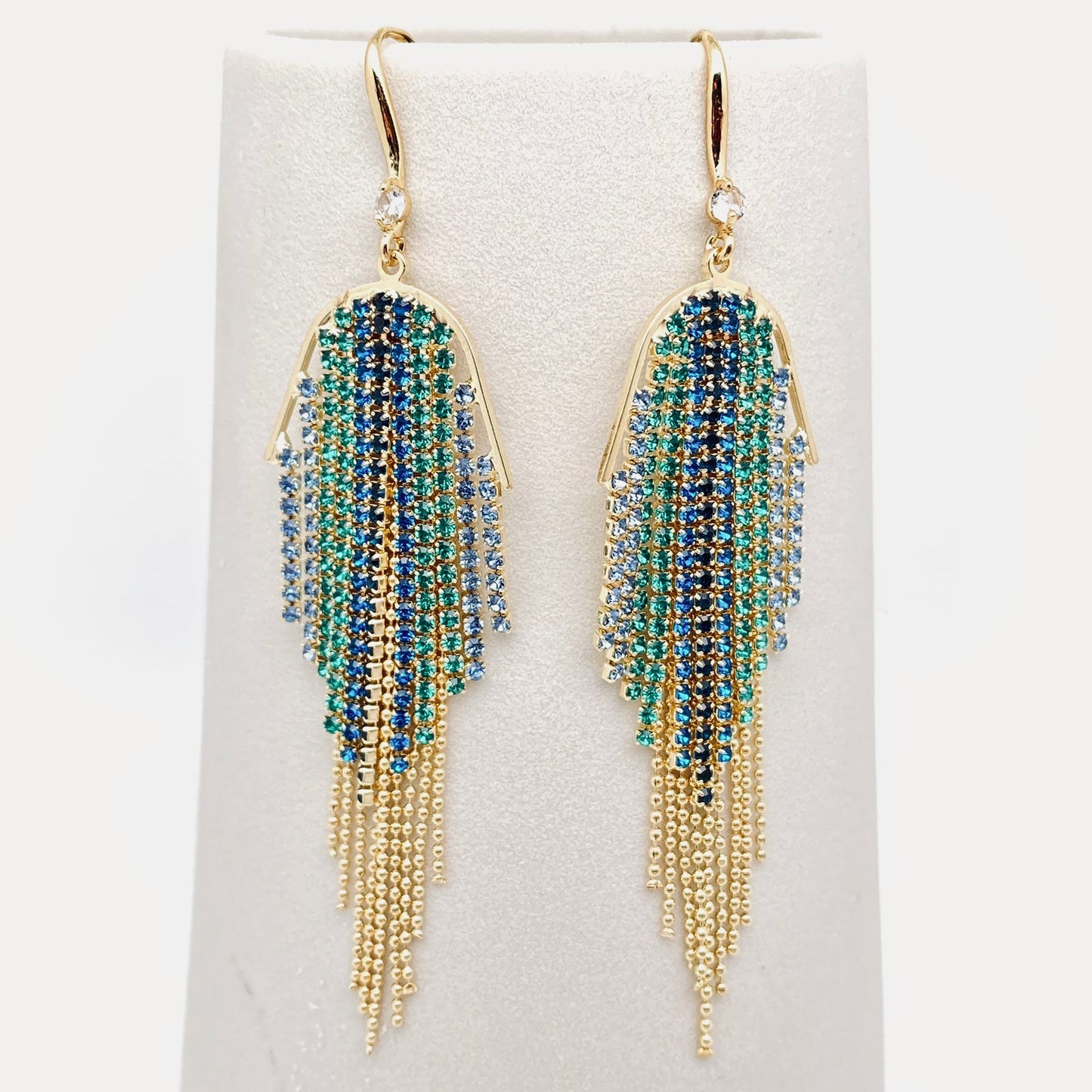 A pair of gold-tone dangle earrings with cascading fringe made of blue, teal, and green rhinestones, displayed on a white jewellery stand.