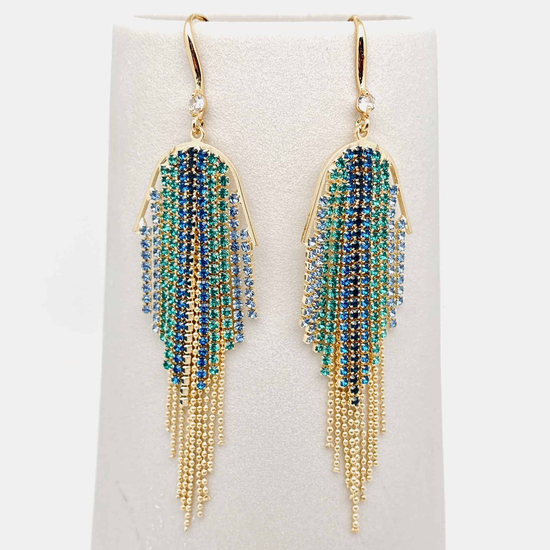 A pair of gold-tone dangle earrings with cascading fringe made of blue, teal, and green rhinestones, displayed on a white jewellery stand.