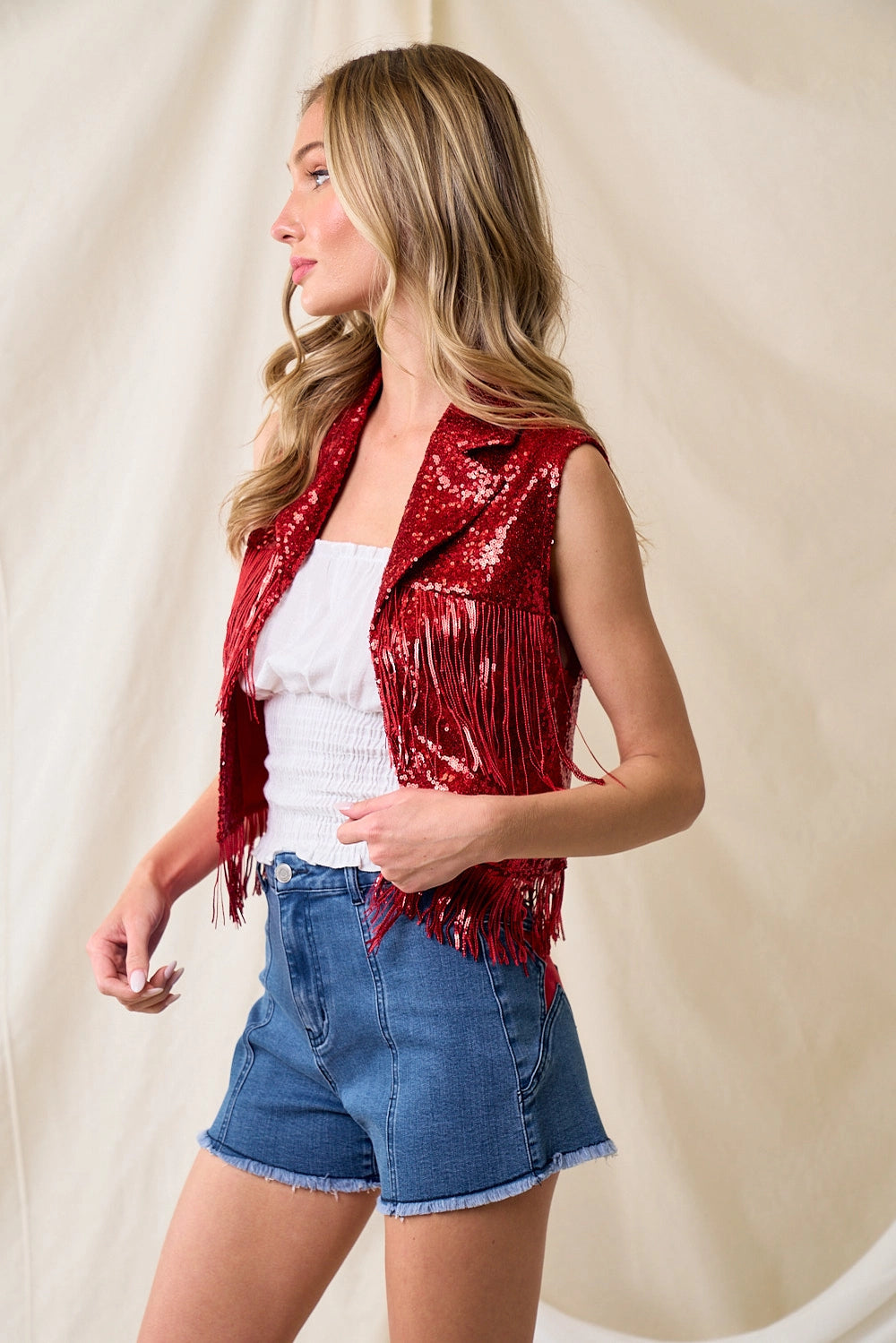 Woman wearing a red sequin vest over a white top and blue shorts on a beige background