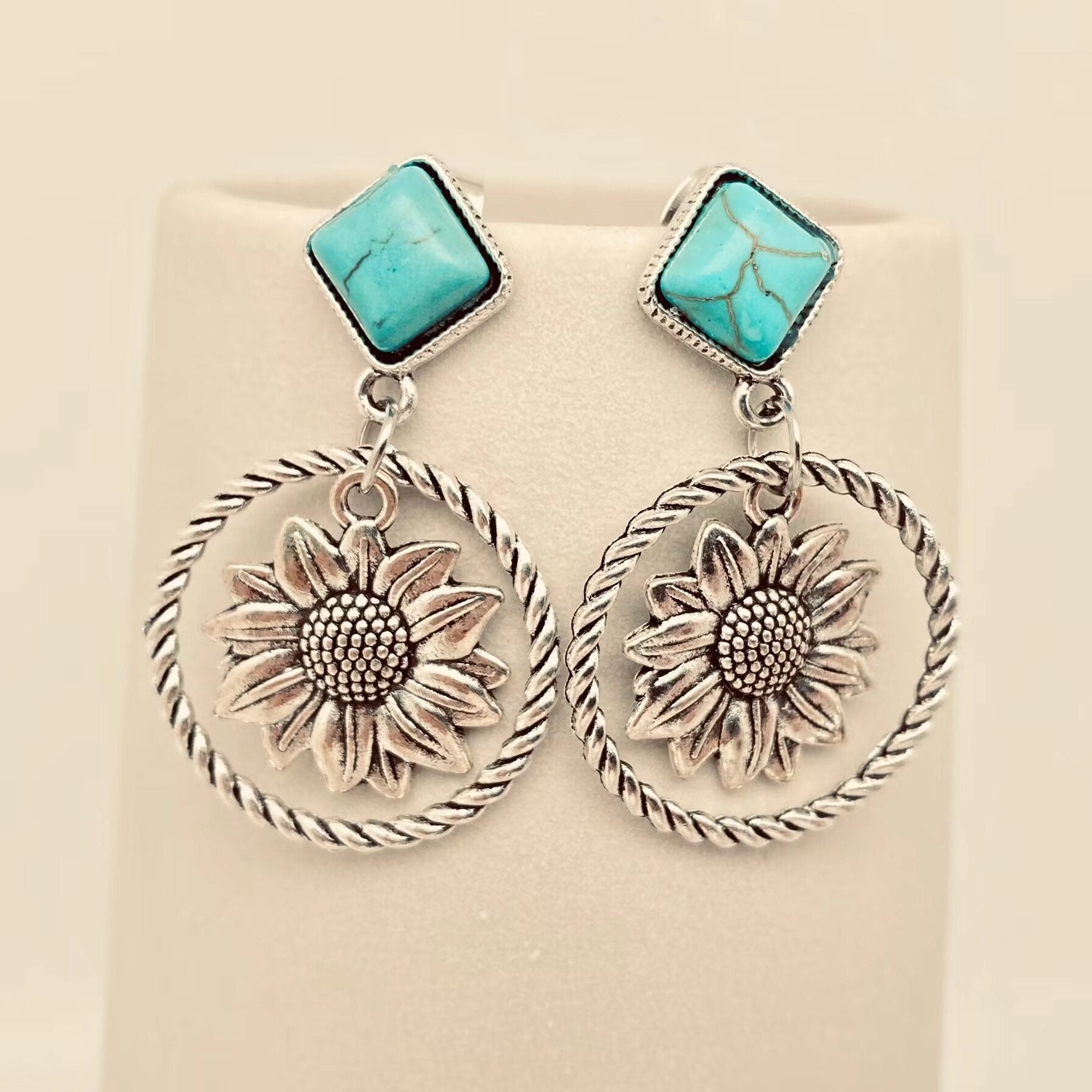 A pair of silver-toned dangle earrings with square turquoise stones at the top and large sunflower motifs inside twisted circular hoops, displayed on a cream jewellery stand.