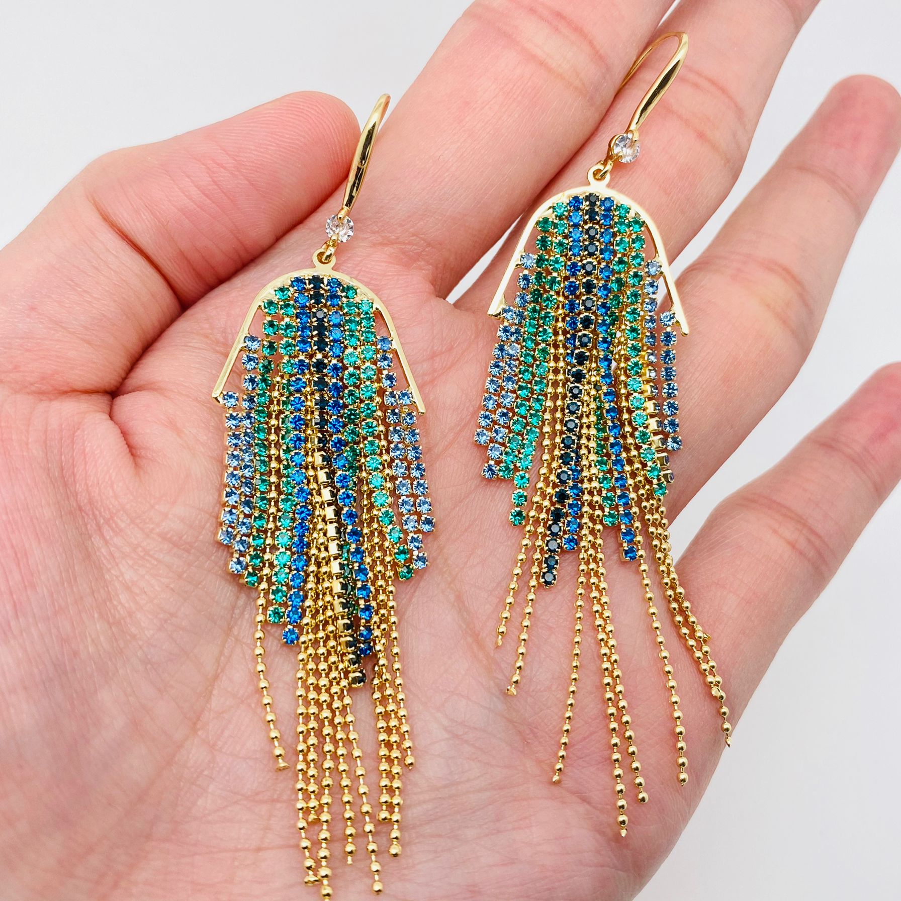 A pair of gold-tone dangle earrings with cascading blue, green, and turquoise rhinestone fringe, held in a person's hand against a light gray background.