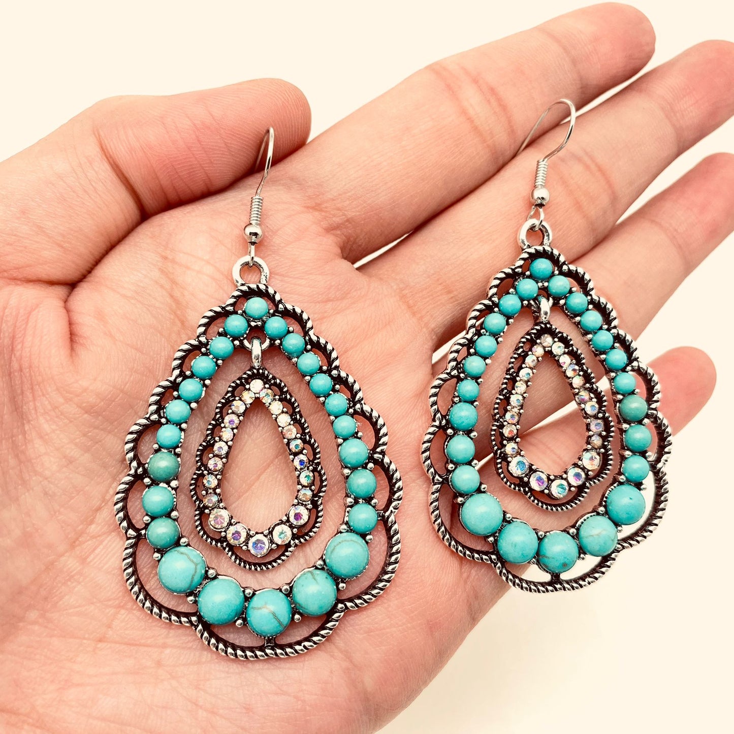 A pair of silver-toned teardrop dangle earrings with turquoise beads and clear rhinestones, held in a person's hand against a light beige background.